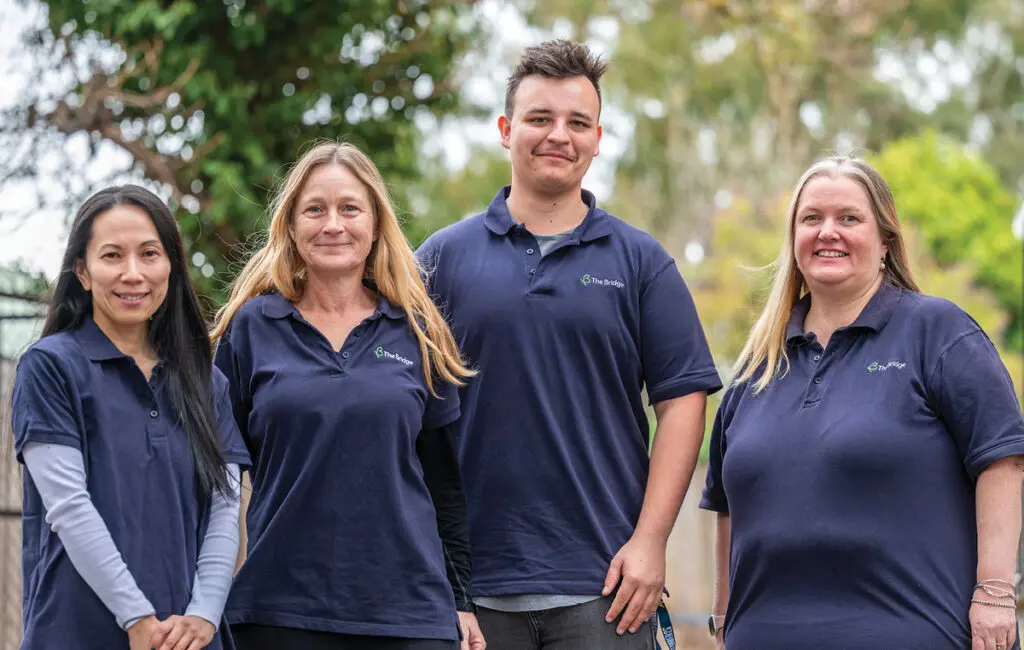 Four people in navy 'The Bridge' polo shirts stand smiling outdoors among trees.