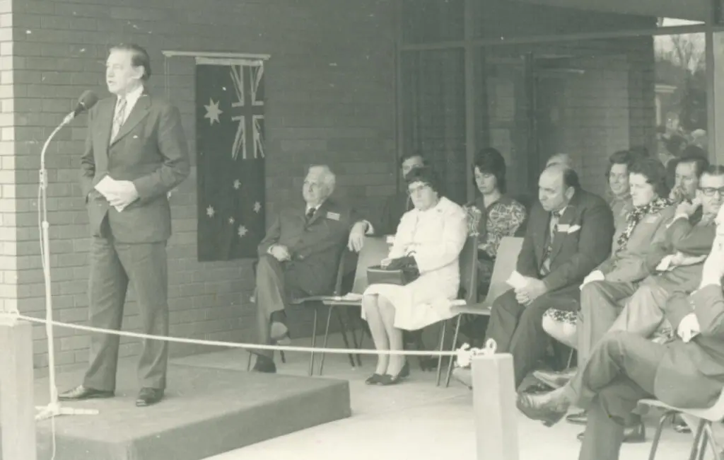 Man in suit speaks at microphone on a small stage, seated audience beside an Australian flag, black and white photo.