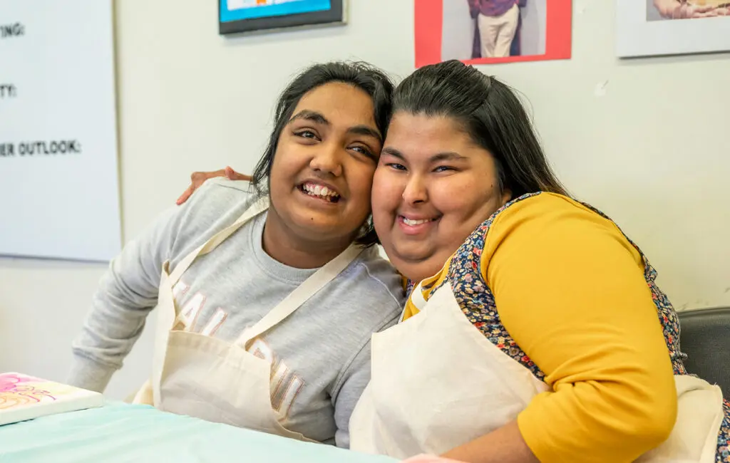 Two women in aprons smile at a table with paintings, artwork displayed on the wall behind them.