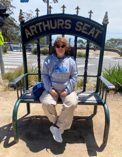 Person in sunglasses and grey hoodie sits on oversized bench labelled 'Arthurs Seat'