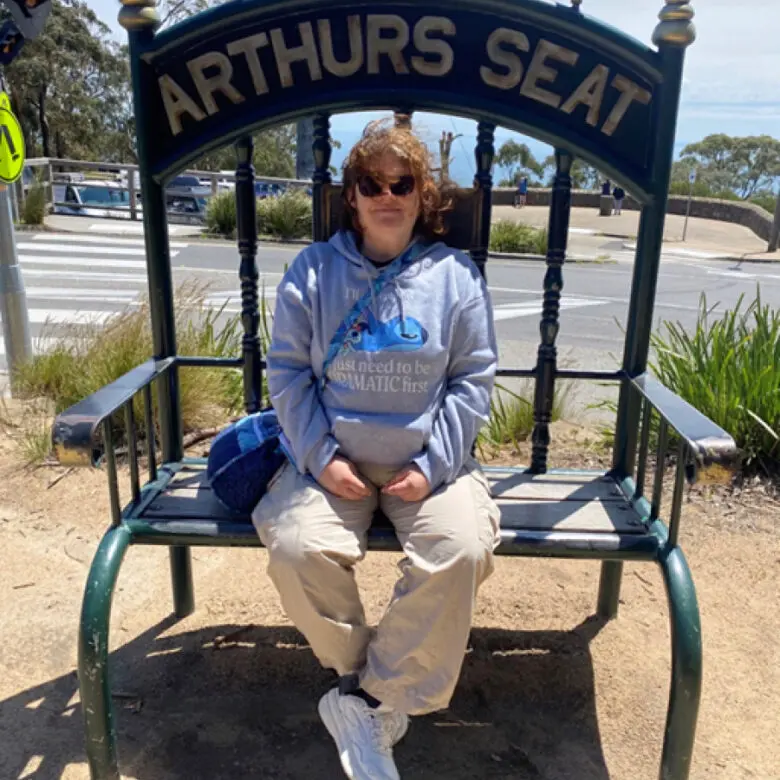 Person in sunglasses and grey hoodie sits on oversized bench labelled 'Arthurs Seat'