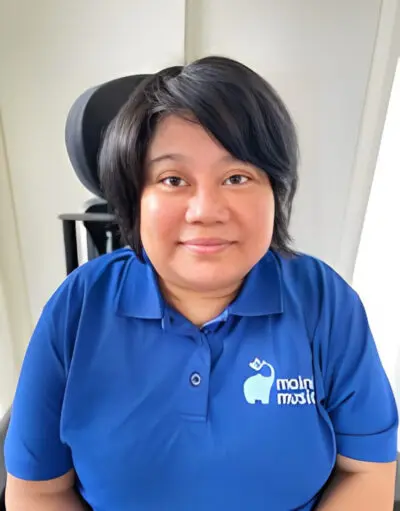 Woman with shoulder-length dark hair smiles, wearing a blue polo shirt with an elephant logo, seated in a wheelchair with headrest.