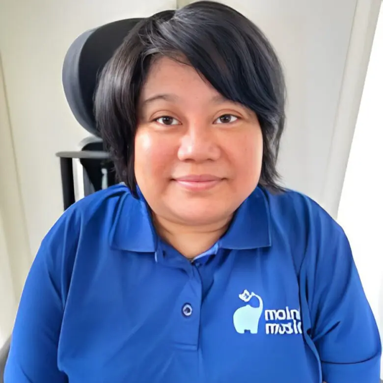 Woman with shoulder-length dark hair smiles, wearing a blue polo shirt with an elephant logo, seated in a wheelchair with headrest.