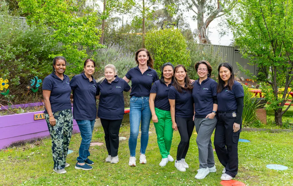 Eight women in matching navy polo shirts stand together smiling in a garden with a raised planter bed.