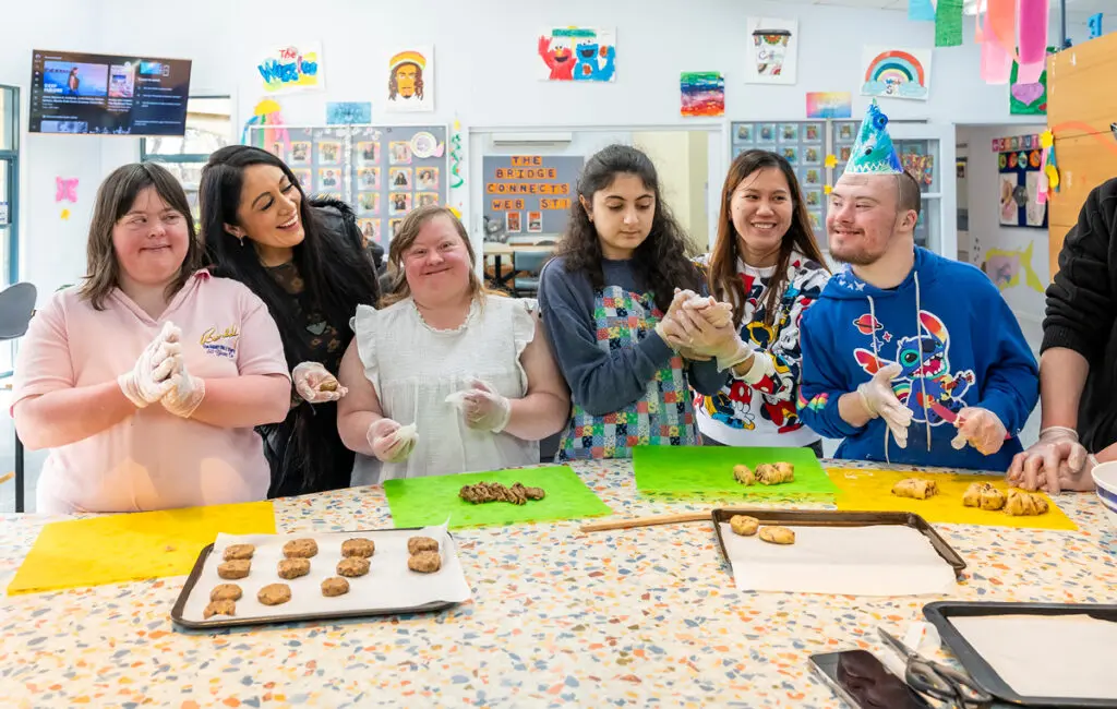 Group wearing gloves shapes cookie dough at a table, smiling; one man wears a party hat, trays hold baked cookies.