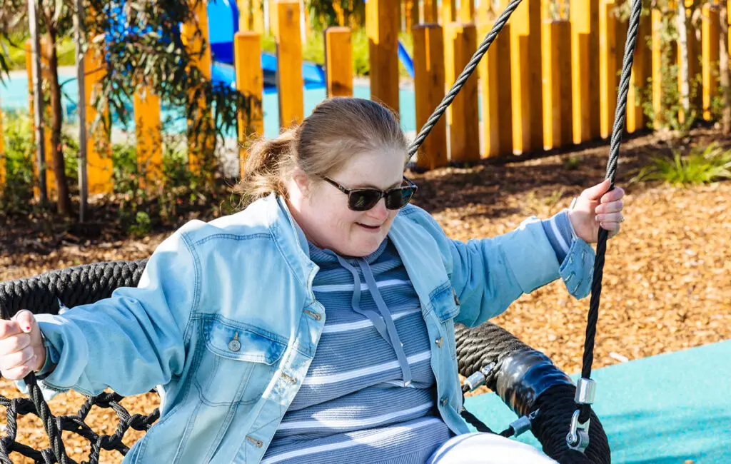 Woman in sunglasses and denim jacket smiling on a rope swing at a playground.