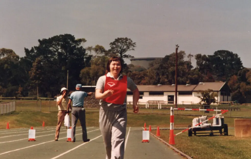 Woman running on a track wearing a red bib, smiling, with two officials and cones nearby.