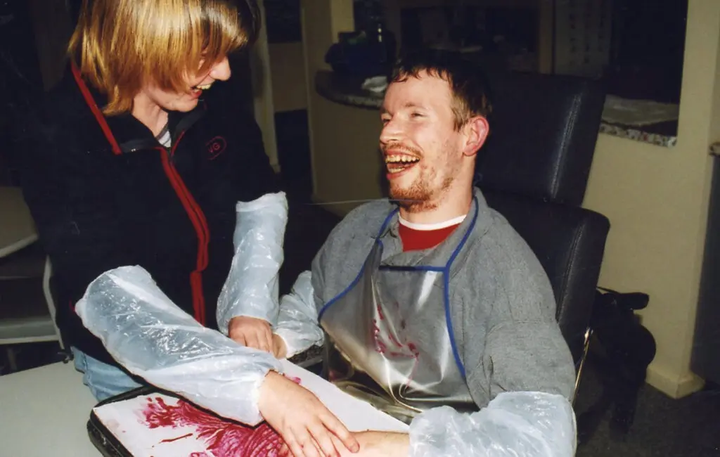 Man in wheelchair laughing while making arm paint art with support worker, both wearing plastic sleeves