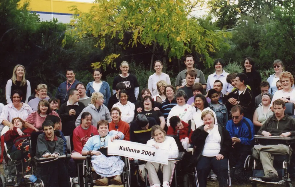 Large group photo outdoors, some in wheelchairs, holding a sign reading 'Kalimna 2004'