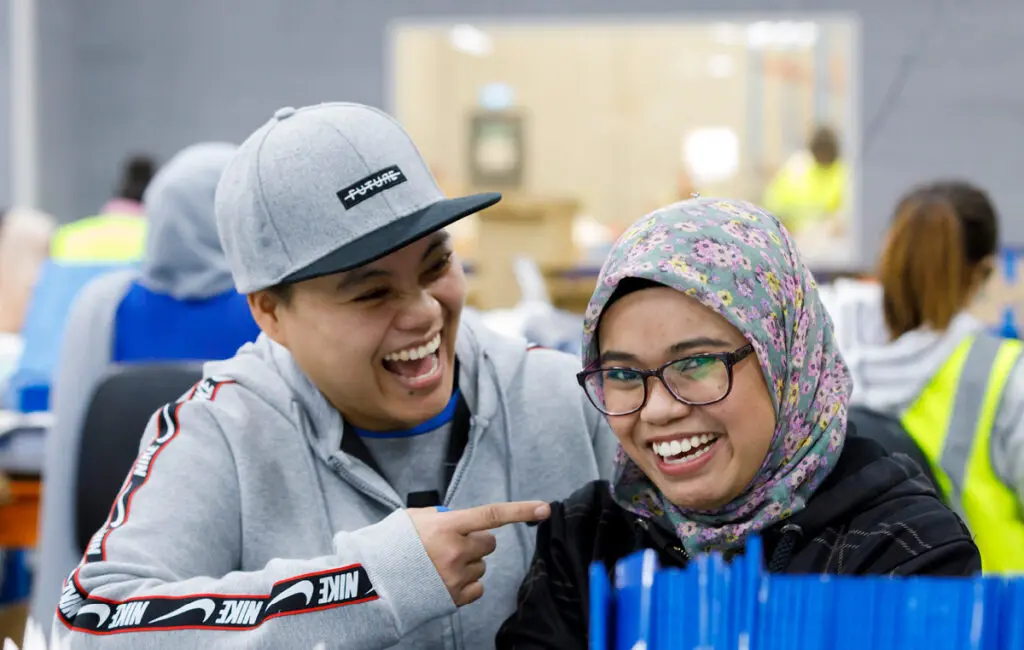 Two smiling workers at a table, one in a cap pointing, the other wearing a floral hijab and glasses, in a workshop.
