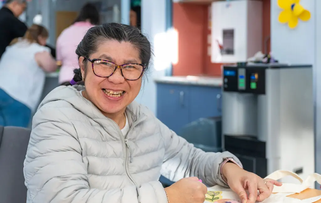 Woman in glasses and grey jacket smiles at camera while decorating a tote bag at a community craft table.