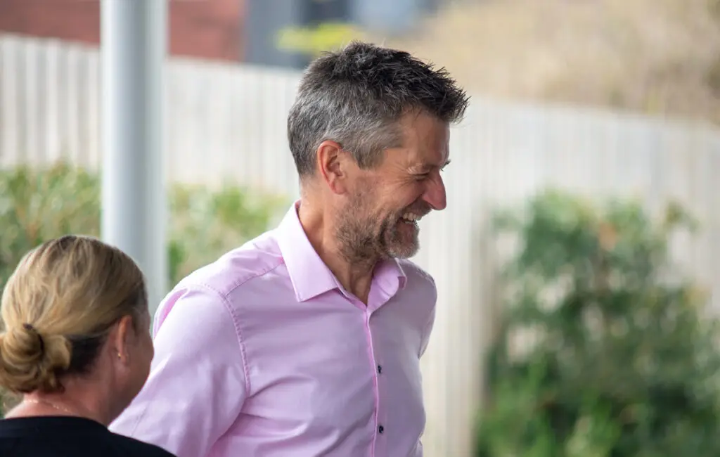 Man in a pink shirt laughing outdoors, standing beside a woman with hair tied back.