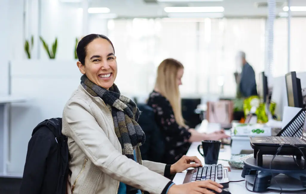 Woman smiling at camera while sitting at a keyboard in an open-plan office, colleagues working at desks behind her