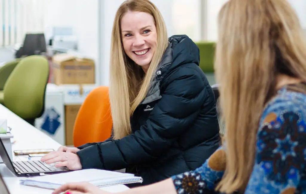 Woman in a puffer jacket smiles while typing on a laptop, seated beside a colleague at an office desk.