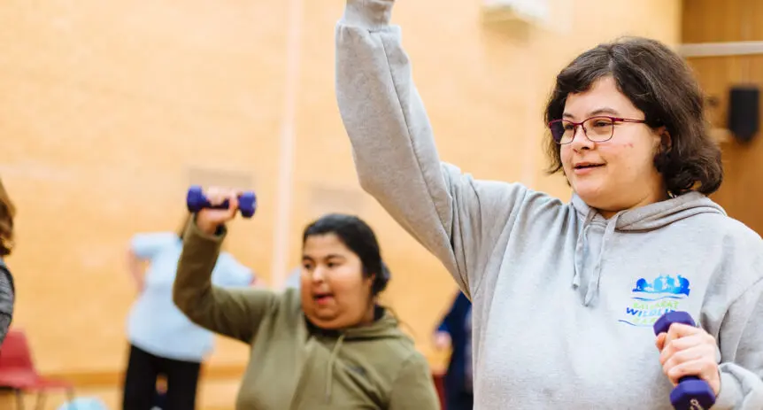 Woman in grey hoodie raises a hand weight overhead during a group exercise class in a hall.