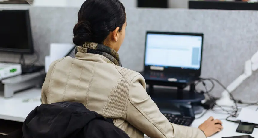 Person seen from behind working at a laptop on a raised stand in an open-plan office.