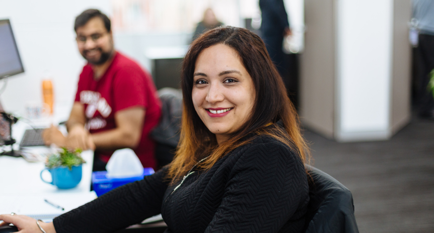 Woman in black cardigan smiling at desk, with a man in a red shirt working behind her in office.