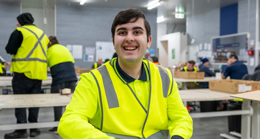 Man in high-visibility vest smiling at camera in a warehouse, with coworkers at tables in the background.