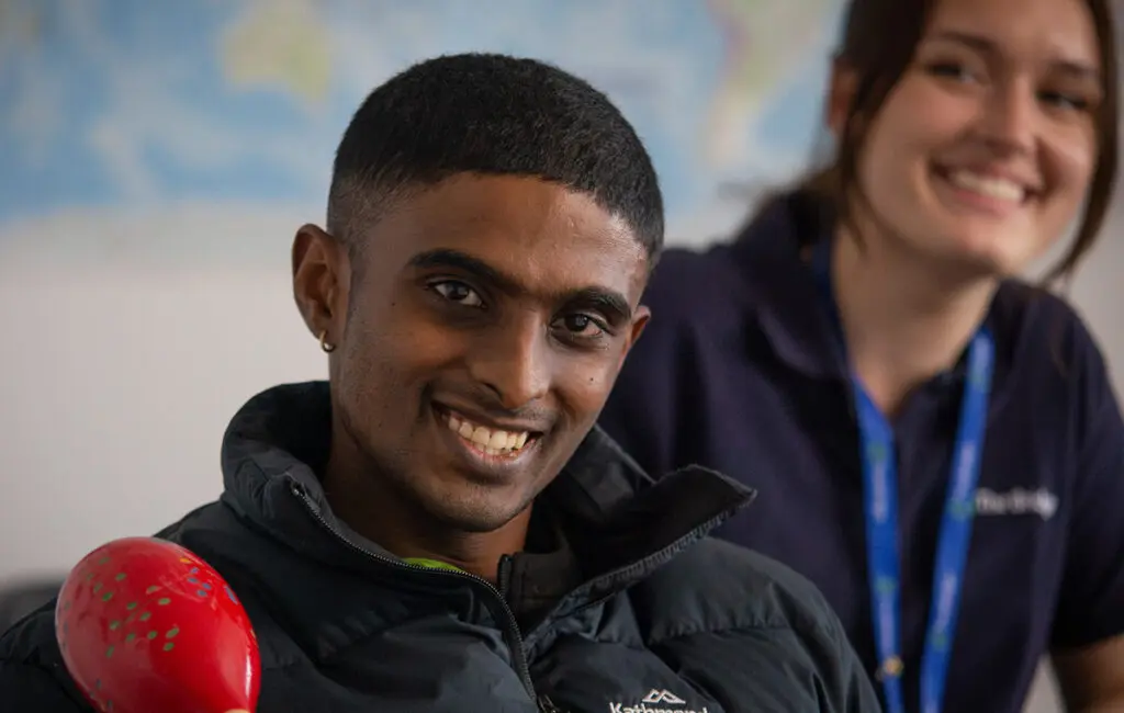 Man smiling, holding a red maraca, with a support worker wearing a lanyard smiling behind him