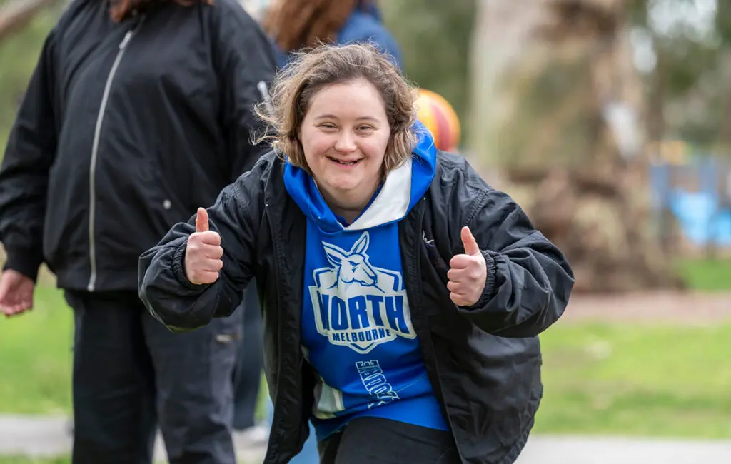 Woman in North Melbourne jersey and jacket smiling, giving two thumbs up outdoors.