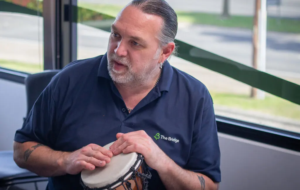 Man in a navy polo shirt with 'The Bridge' logo playing a djembe drum indoors near a window.