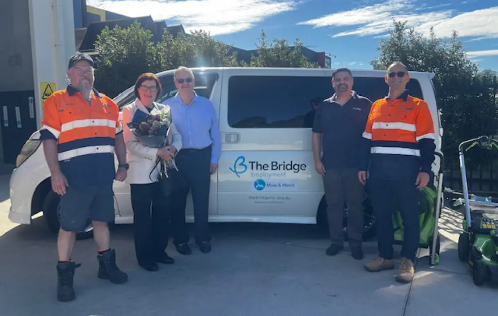 Five people stand beside a white van reading The Bridge Employment Mow & Mend, woman holding flowers