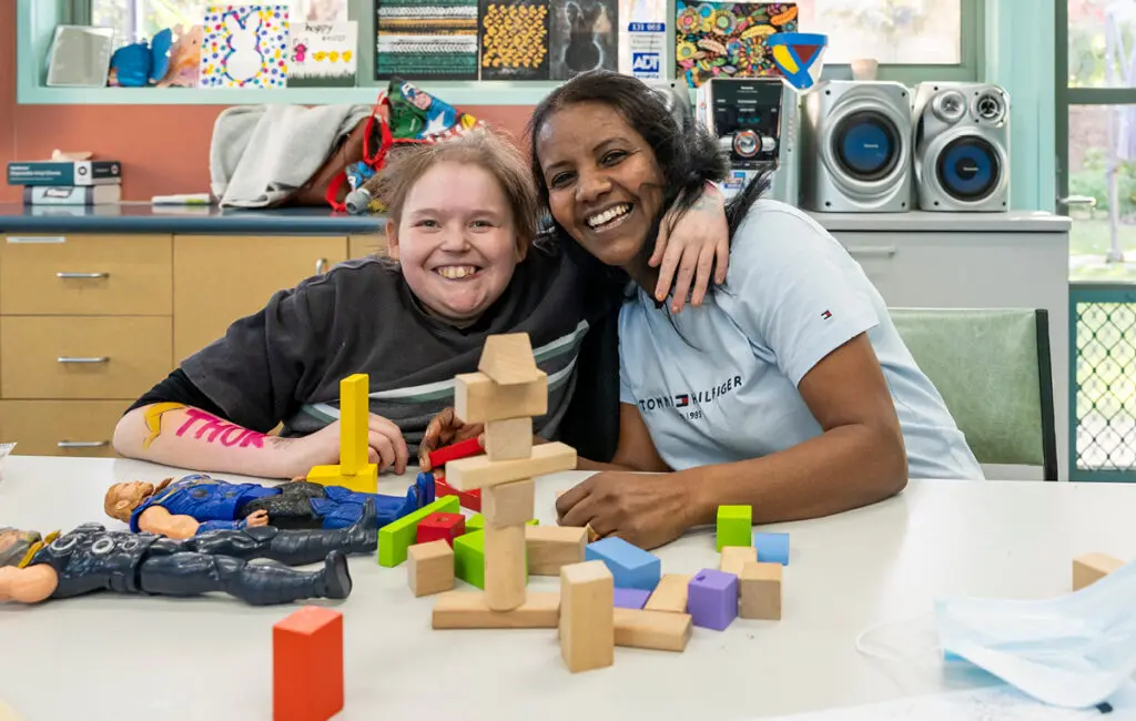 A support worker and a young person smile at a table with wooden blocks and action figures in a community room.