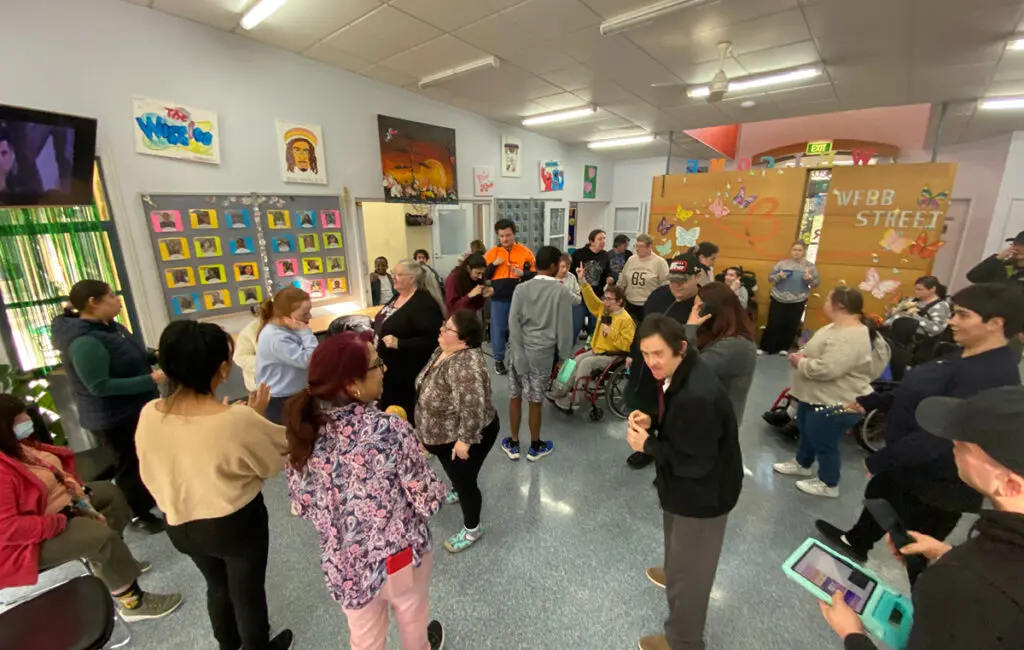 Group gathers in a community room decorated with art and a 'Webb Street' sign, some standing, some using wheelchairs