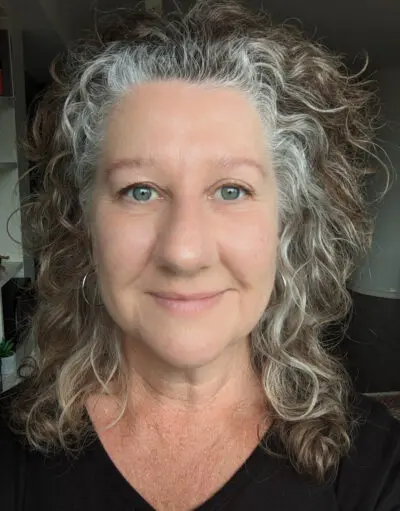 Woman with curly grey and brown hair, hoop earrings, wearing a black top, smiling indoors.