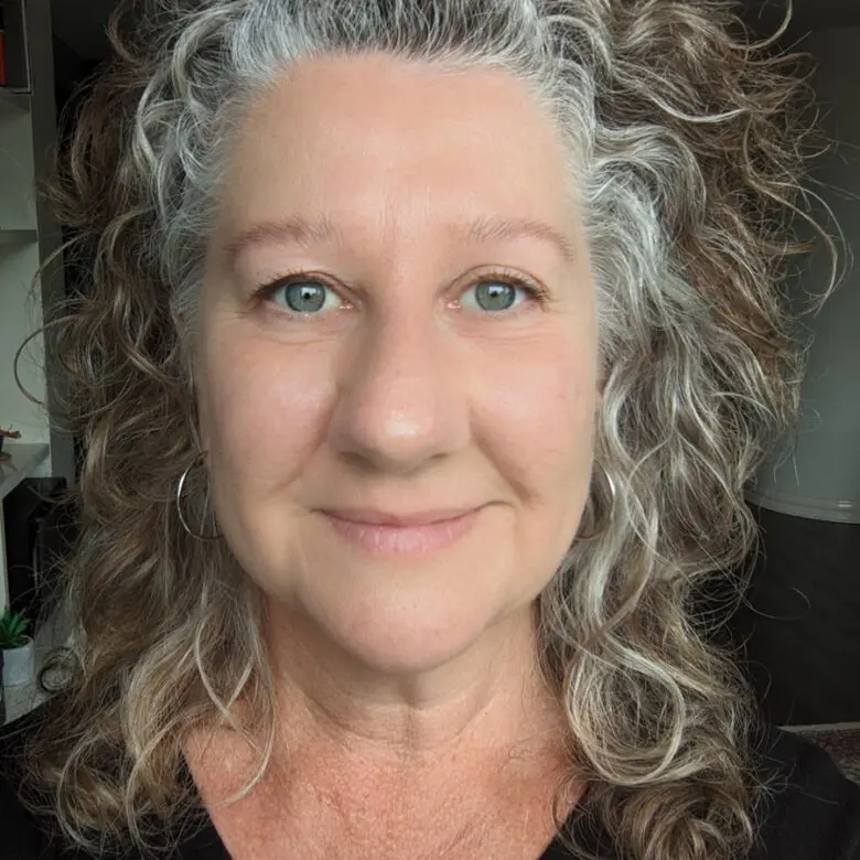 Woman with curly grey and brown hair, hoop earrings, wearing a black top, smiling indoors.