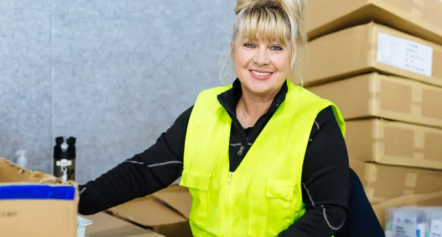 Woman in a high-visibility vest smiles beside stacked cardboard boxes and packing supplies.
