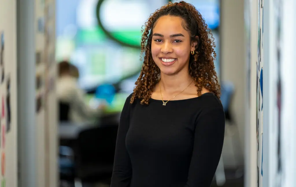 Woman with curly hair smiling in a hallway, wearing a black top, office space blurred behind her.