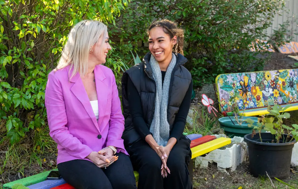 Two smiling women sit talking on a colourful painted bench in a garden with a mosaic bench nearby.