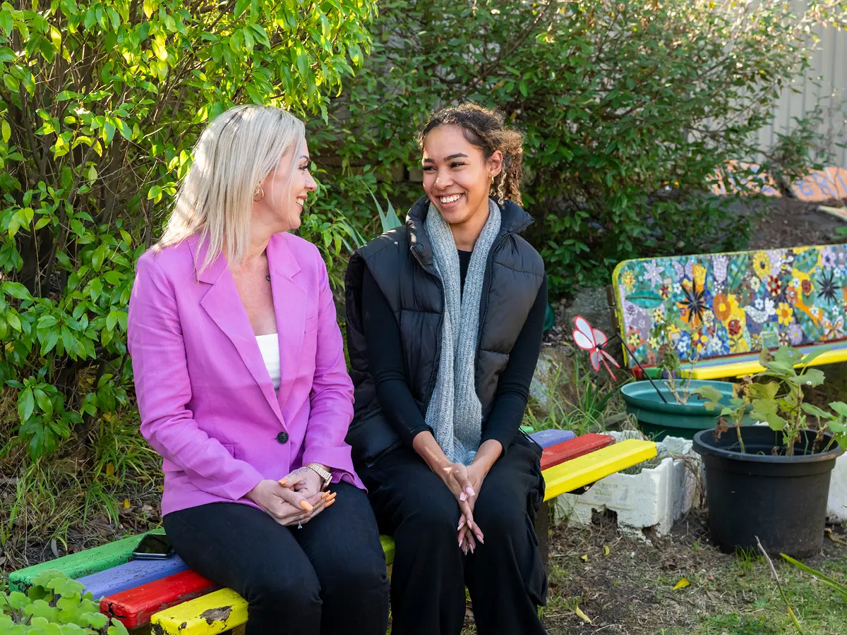 Two smiling women sit talking on a colourful painted bench in a garden with a mosaic bench nearby.