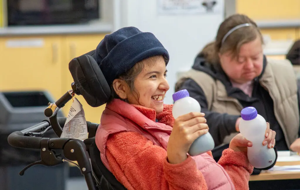 Smiling woman in wheelchair with headrest holds a small bottle; another person sits nearby indoors.