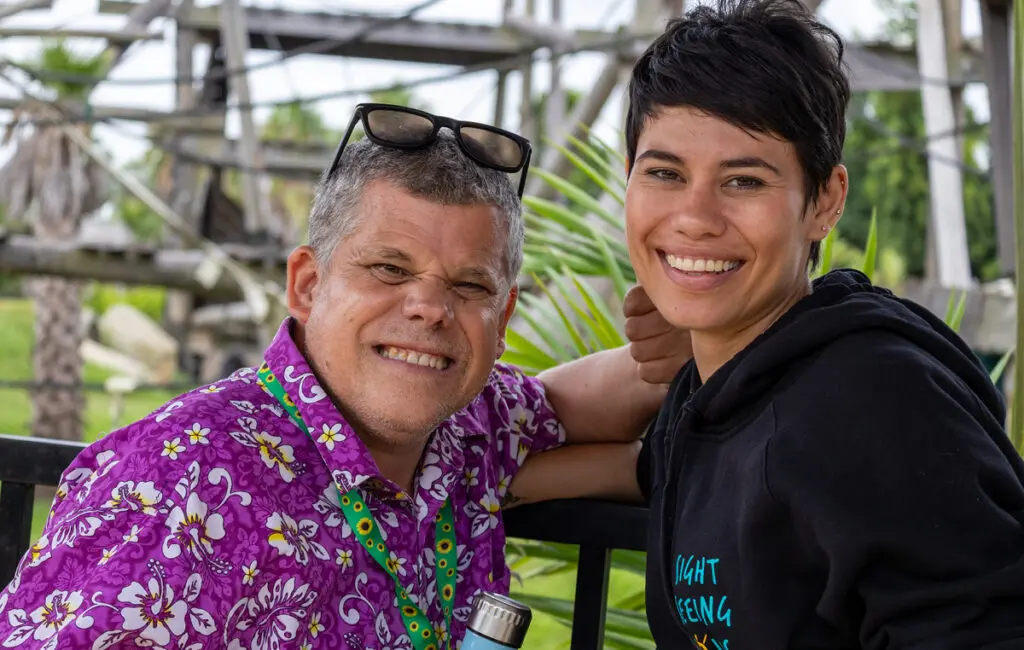 Man in a purple floral shirt and woman in a black hoodie smiling together outdoors near a play structure.