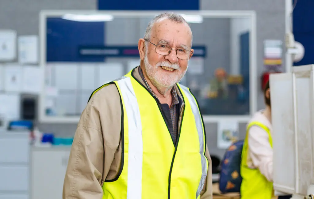 Man with beard and glasses smiles wearing a hi-vis vest, with another worker in the background.