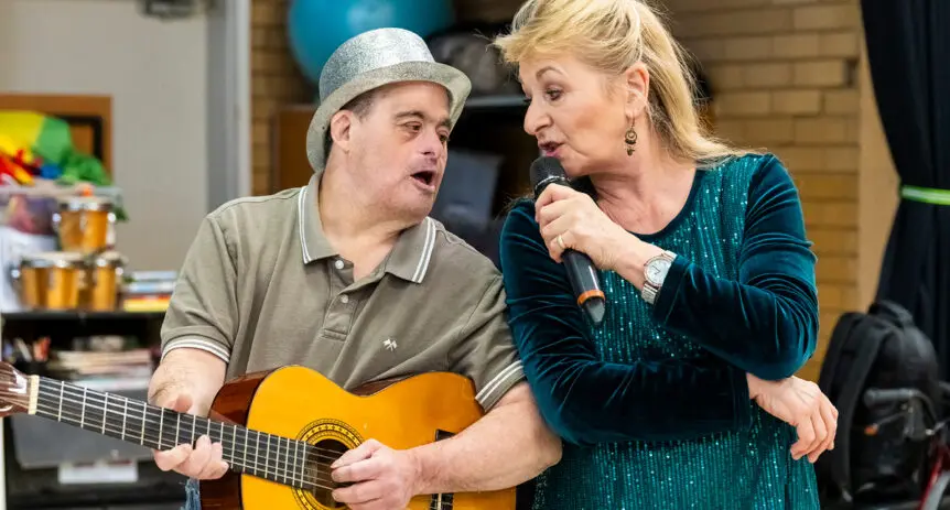 Man in a silver hat plays guitar and sings beside a woman singing into a microphone