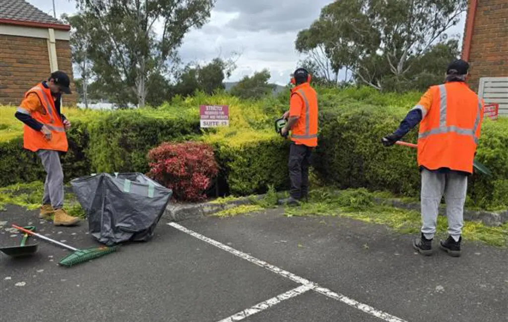 Three workers in orange hi-vis vests trim and rake a hedge in a car park, with a collapsible waste bag nearby.