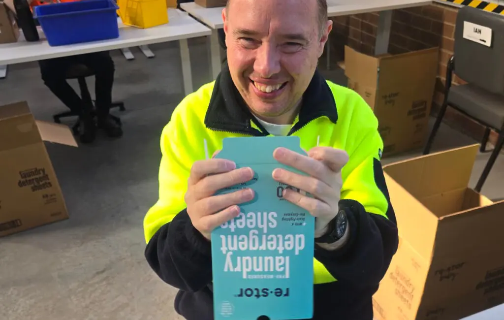 Man in a yellow safety jacket smiles, holding a box of laundry detergent sheets in a warehouse workspace.