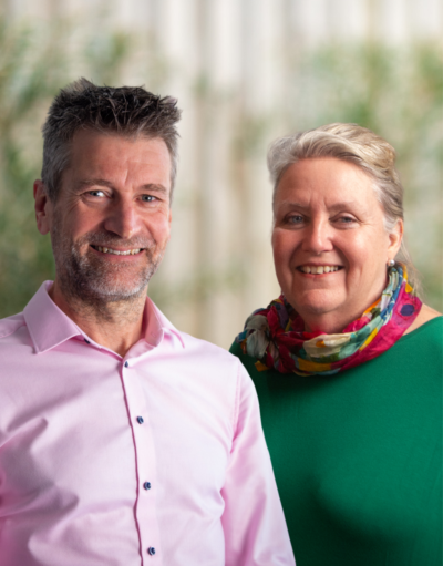 A man in a pink shirt and a woman in a green top with a floral scarf smile at the camera.