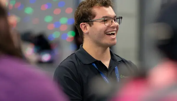Man with curly hair and glasses laughs, wearing a dark polo shirt and lanyard, with people blurred in foreground.