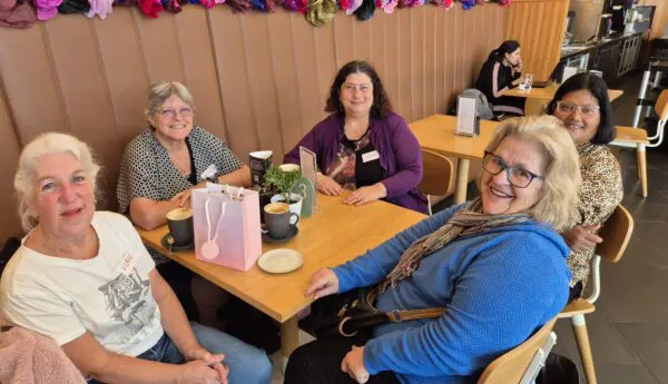 Five women smiling at a cafe table with coffees and a gift bag, fabric flower garland on wall behind.