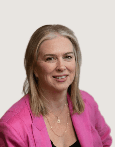 Woman with shoulder-length blonde hair, smiling, wearing a pink blazer and necklace, against a light background.