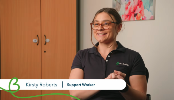 Kirsty Roberts, Support Worker, smiling in a navy The Bridge polo shirt, seated indoors near a wardrobe.