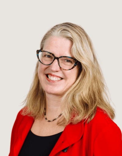 Woman with long blonde hair and patterned glasses, smiling, wearing a red blazer and beaded necklace.
