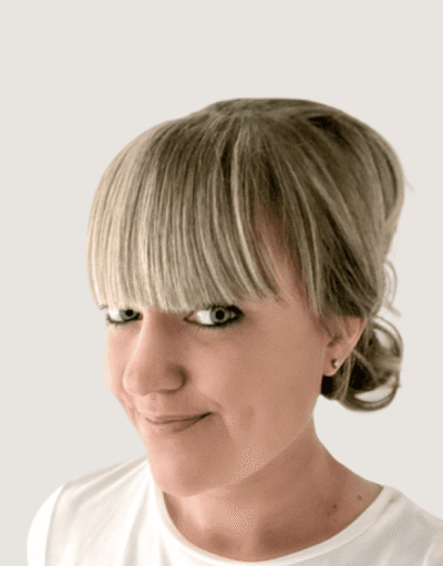 Close-up portrait of a woman with blunt fringe and hair tied back, wearing a white top, smiling slightly.