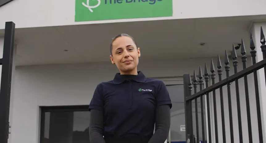 Woman in The Bridge uniform stands by a metal gate outside the building, with The Bridge sign above the entrance.