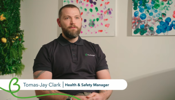 Tomas-Jay Clark, Health & Safety Manager, seated in The Bridge polo shirt, with colourful artwork behind him.