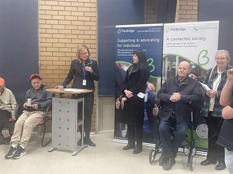 Woman speaking at a lectern beside two banners for The Bridge, with seated audience and standing attendees nearby.